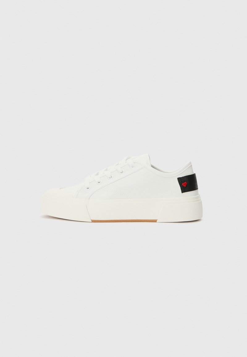 Sneaker de lona branca com biqueira redonda, sola de borracha plana e uma aba preta no calcanhar com um emblema de coração vermelho. Ilhós para os atacadores na parte da frente.