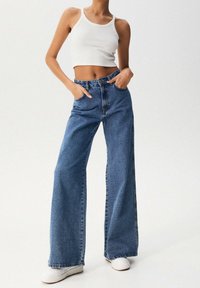 Vaqueros de pierna ancha azules con cintura alta, hechos de denim. Presentan bolsillos frontales, un cierre de botón y un desvanecimiento sutil. Combinados con un top corto blanco.