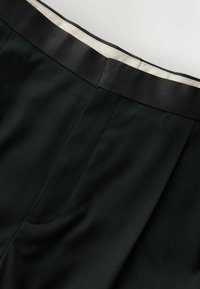 Čierne na mieru šité nohavice z hladkej látky s bielym kontrastným detailom na páse, zobrazené spredu v oblasti pása.