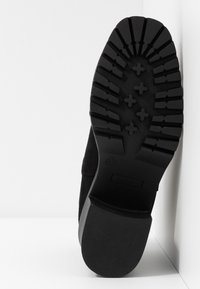 Semelle noire d'une botte avec des motifs de bande de roulement profonds, présentant des crampons en forme de croix et un talon bloc, photographiée sur un fond blanc.