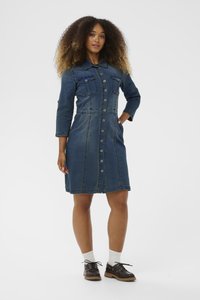 Kvinna med lockigt hår som bär en knäppt denimklänning, vita strumpor och mörkbruna snörskor, stående med ena handen i fickan.