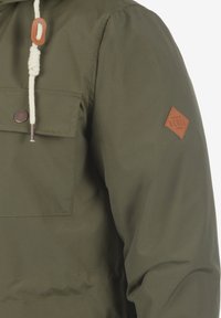 Olivenfarbene Jacke mit einer großen Fronttasche, beigem Kordelzug mit Holzstopper und einem Lederpatch mit der Aufschrift "Rebel" am Ärmel.