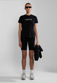 Black cotton t-shirt with logo, paired with black leggings. White socks and light sneakers complete the outfit. Holding a black jacket.