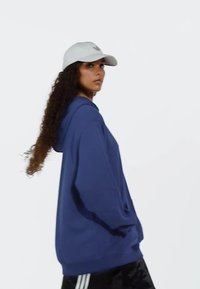 Blå hoodie med en framficka och dragsnodd på huvan, tillsammans med en vit keps. Modellen har lockigt hår och visas från sidan mot en neutral bakgrund.