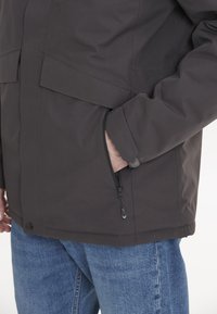 Mörkgrå jacka med en dragkedjeficka, knappstängning och strukturerad tyg. Bärs med blåjeans, vilket visar en avslappnad stil.