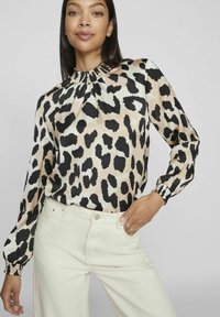 Blus med leopardmönster i beige och svart, med en gathered-hals, långa ärmar med elastiska manschetter och en silkesliknande textur. Matchad med vita byxor.