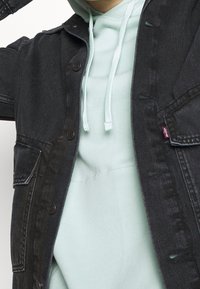 Svart denimjacka med bröstfickor och knappstängning, lager-på-lager över en ljus mintgrön hoodie med dragsko. Mjuka texturer är tydliga.