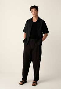 Black short-sleeve shirt with a textured pattern, paired with loose black trousers and black sandals, featuring a relaxed fit and modern design.