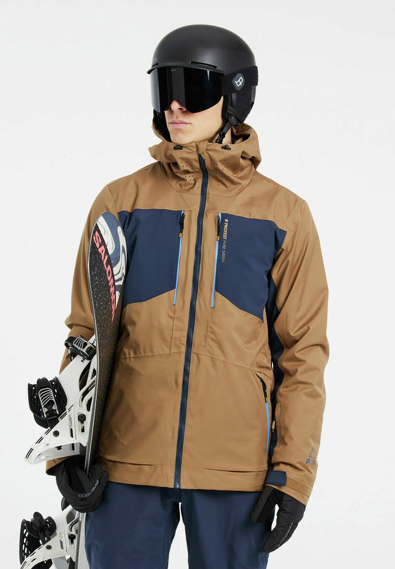 Brun och marinblå skidjacka med huva, två zipfickor och blåa dragkedjor. Svarta handskar och ett snowboard med vita bindningar fulländar looken.