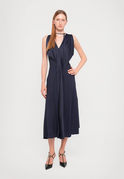 Laivansinis sleeveless mekko, jossa on V-pääntie, drapattu muotoilu, vyötärösidokset ja laskeutuva hame. Yhdistetty mustiin teräväkärkisiin korkokenkiin ja nilkkaketjuihin.