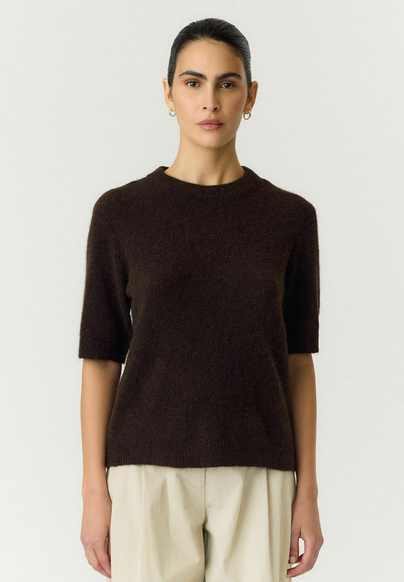 Femme aux cheveux foncés tirés en arrière portant un pull à manches courtes marron foncé et un pantalon beige clair, debout devant un fond uni.