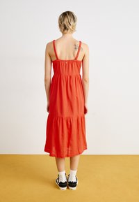Robe midi rouge fluide avec des bretelles volantes et un design à étages. Portez-la avec des baskets noires et des chaussettes blanches, sur un sol jaune.