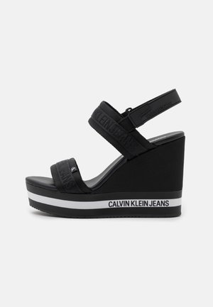 Czarna sandałka na koturnie z dwoma regulowanymi paskami z tkaniny i logo Calvin Klein Jeans na podeszwie oraz paskach, widok z boku na białym tle.