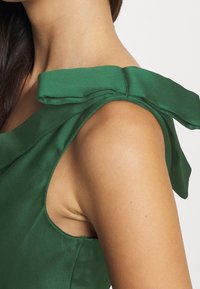 Nainen, joka käyttää vihreää hihattomaa toppia, jossa on kangaspainatus olalla. Lähikuva kaulasta ja yläkädestä.