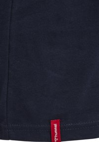 Mörkblå bomullströja med en böjd fåll och en kontrasterande röd etikett i nederkanten. Slät textur, minimalistisk design.