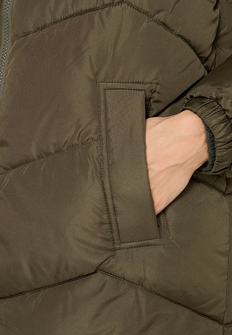 Hand teilweise in der vorderen Tasche einer dunkelolivgrünen gepolsterten Winterjacke mit elastischen Bündchen.