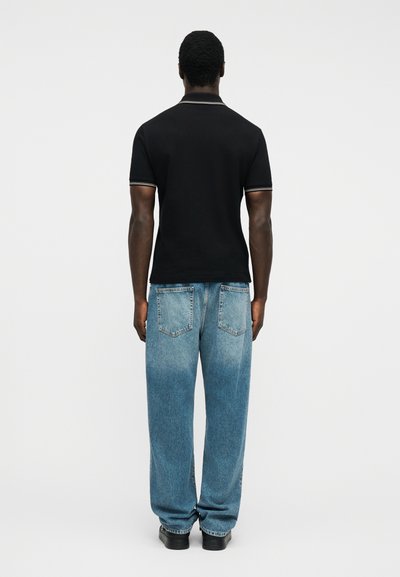 Άνδρας που στέκεται με την πλάτη στραμμένη, φορώντας μαύρο κοντομάνικο πόλο και άνετο μπλε τζιν με μαύρα παπούτσια, μπροστά σε απλό φόντο.