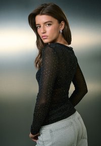 Melnas caurspīdīgas garām piedurknēm blūze ar metalizētajiem akcentiem, pieguļošs siluets un teksturēts audums. Apvienota ar gaiši pelēkiem, augsti novietotiem džinsiem.