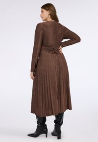 Lång, bronsfärgad plisserad klänning med långa ärmar, figursydd midja och markerad rygg, kombinerad med svarta ankelstövlar. Slätt, texturerat tyg.