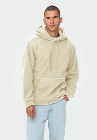Flauschiger beiger Hoodie mit einer Kordelzugkapuze, Fronttasche und strukturiertem Stoff; kombiniert mit hellblauer Jeans.