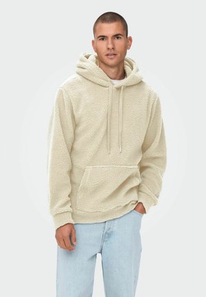 Fuzzy beige hoodie met een trekkoordkap, een voorzak en een textuur stof; gecombineerd met lichtblauwe jeans.
