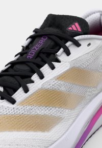 Scarpa sportiva con una tomaia in rete bianca e oro, lacci neri e accenti viola. Presenta una suola texturizzata e un prominente design a tre strisce.