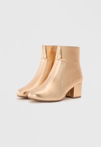 Botas de tobillo metálicas doradas con un diseño elegante, textura suave, punta afilada y tacón cuadrado. Presenta un detalle de costura lateral.