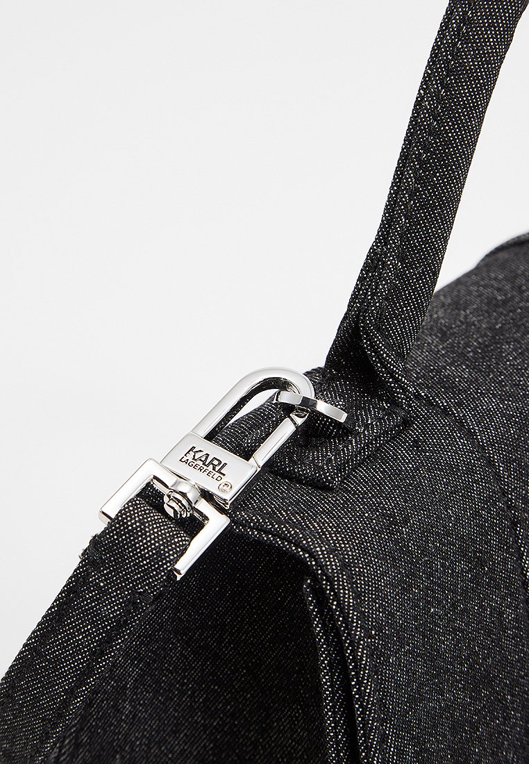 Gros plan sur un coin de sac en tissu noir texturé avec un fermoir en argent gravé "KARL LAGERFELD" reliant la sangle au sac.