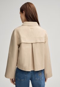 Giacca corta beige con retro plissettato, dettaglio del logo dorato al colletto e maniche lunghe, abbinata a jeans blu con logo ricamato.