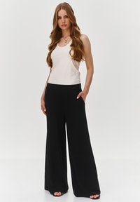Kremowy top na ramiączkach w połączeniu z szerokimi, czarnymi spodniami. Ubranie ma dopasowany krój w talii oraz kieszenie, wykonane jest z gładkiego, lekkiego materiału.