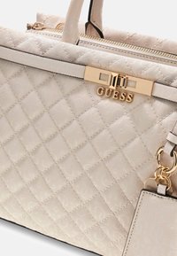 Gep quiltete cremefarbene Handtasche mit goldfarbenem GUESS-Logo, ausgestattet mit einem Reißverschluss, einem Kettenakzent und einer strukturierten Oberfläche mit dezentem Muster.
