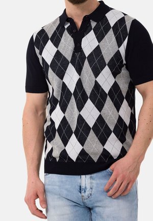 Homme portant un polo à manches courtes en argyle noir, gris et blanc, et un jean bleu clair, main reposant près de la poche.