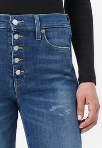 Jeans in denim blu a vita alta con cinque bottoni argentati, una tasca frontale e lievi segni di usura nella zona della coscia. Il materiale ha una texture liscia.