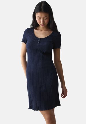 Junge Frau trägt ein marineblau geripptes Kurzarmkleid mit Knöpfen, steht und blickt nach unten vor einem einfachen hellen Hintergrund.