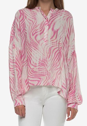 Femme portant un chemisier ample à manches longues rayé zèbre rose et blanc, associé à un pantalon blanc, debout devant un fond uni.