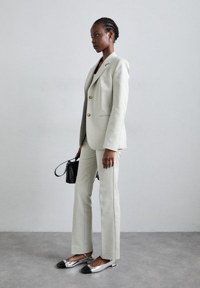 Hellegrau maßgeschneiderter Anzug mit goldenen Knöpfen, kombiniert mit metallischen Schuhen mit schwarzen Zehenakzenten. Eine kleine schwarze Handtasche rundet das Outfit ab.