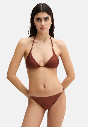 Kobieta o długich ciemnych włosach, ubrana w brązowy dwuczęściowy bikini z koralikowymi ramiączkami, stojąca z rękami za plecami na jednolitym tle.