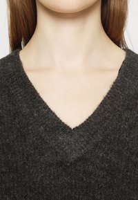 Maglione grigio scuro con scollo a V, realizzato in un tessuto a maglia testurizzato, caratterizzato da una leggera morbidezza e dettagli a bordo irregolare attorno al collo.