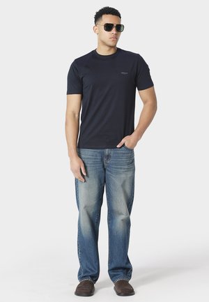 Mann trägt schwarze Sonnenbrille, ein dunkelblaues kurzärmeliges T-Shirt, ausgebleichte blaue Jeans und braune Schuhe, steht mit einer Hand in der Hosentasche.