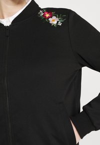 Černá bomber bunda na zip s květinovou výšivkou v červených, růžových a zelených tónech na levém rameni. Měkká, strukturovaná tkanina.