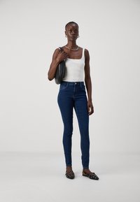 Ψηλόμεσα σκούρα μπλε skinny τζιν, συνδυασμένα με λευκή ριγέ αμάνικη μπλούζα και μαύρες φλατ παπούτσια με καρφιά. Το μοντέλο κρατά μια μαύρη στρογγυλή τσάντα.