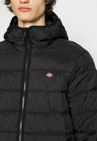 Black puffer jacket with a hood, featuring a zip closure and horizontal quilted design. Small logo in red and yellow on the chest.