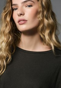 T-shirt verde scuro leggera con scollo rotondo. Il modello ha capelli lunghi, ondulati e biondi. Pelle liscia e trucco minimalista.