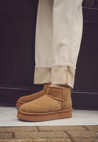 Brune ruskindsankelstøvler med en tyk gummisål, med en rundet snude og en kort skaft, kombineret med lyse bukser med opbukket kant.