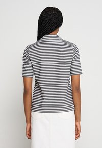 Femme aux cheveux tressés portant un t-shirt à manches courtes rayé gris et blanc et une jupe blanche, debout dos à la caméra.