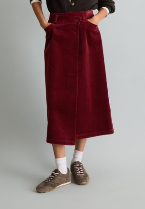 Persoon die een bordeauxrode corduroy rok tot op de knie draagt met zakken, witte sokken en bruine vetersneakers, staand op een lichtgrijze vloer.