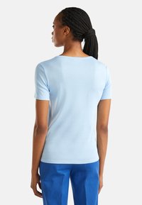 Žena s copy, uvázanými v culíku, oblečená v krátkém světle modrém topu a modrých kalhotách, stojící s dokonale otočeným zády k posluchačům.