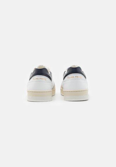 Pár bílých tenisek s námořnickými akcenty na patě a béžovými podrážkami, pohled zezadu, který ukazuje logo "Filling Pieces" ve zlatém písmu.