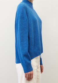 Pull en maille bleu avec un col montant, une coupe décontractée et un ourlet côtelé. Texture lisse et manches longues. Porté sur un pantalon de couleur claire.