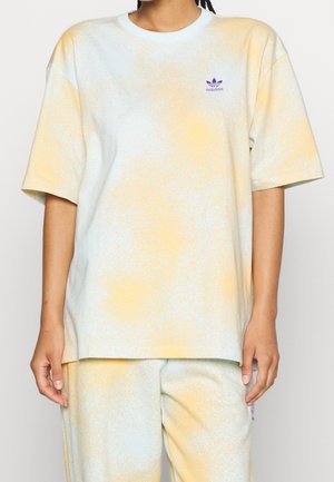 Person iført en løs, kortærmet, tie-dye Adidas t-shirt i gul og lys blå samt matchende bukser med lilla logo på brystet og lår.